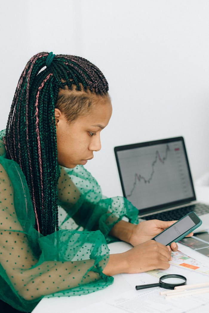Focused woman analyzing financial graphs on laptop and phone for online work.